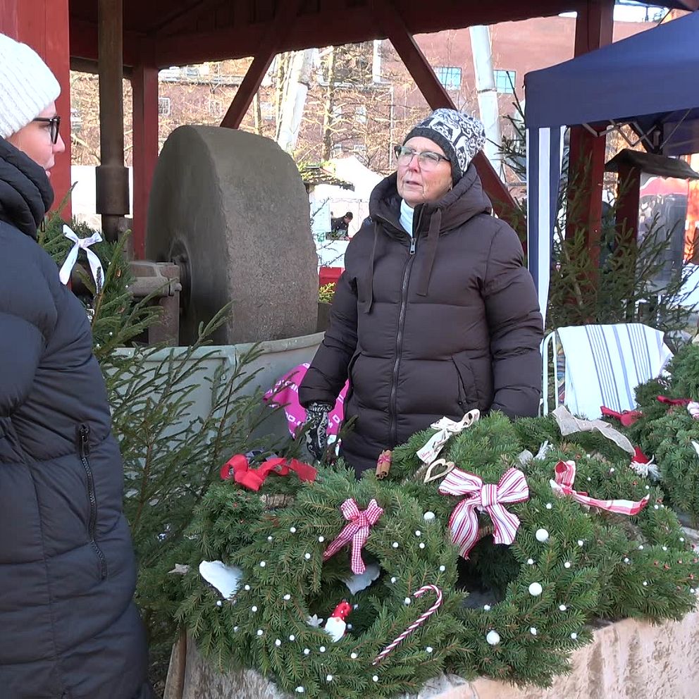 Två personer står vid ett försäljningsstånd som visar upp julkransar dekorerade med rosetter och godis. Ståndet är täckt med en tygduk och omgiven av grönska, inom en utomhusmiljö. I bakgrunden syns en stor sten och andra personer är delaktiga i aktiviteten.