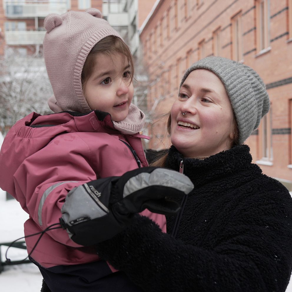 En person bär ett barn i en snötäckt innergård, omgiven av byggnader med murade fasader. Både vuxna och barn är klädda i varma vinterkläder, och snön täcker marken och bänkarna i bakgrunden.