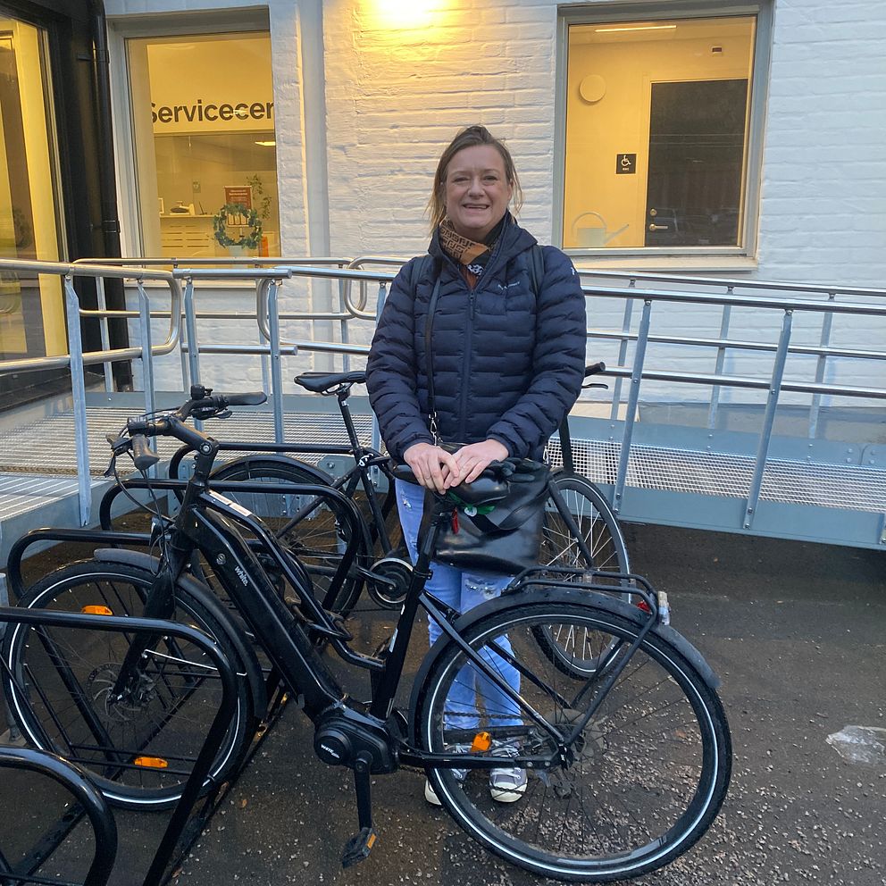 Kajsa Stenlund som är anställd på Sundsvalls kommun står vid ett cykelställ med flera svarta cyklar. Personen bär en mörk jacka och ser ut att vänta. Bakgrunden visar en byggnad med stora fönster och ordet Servicecenter.