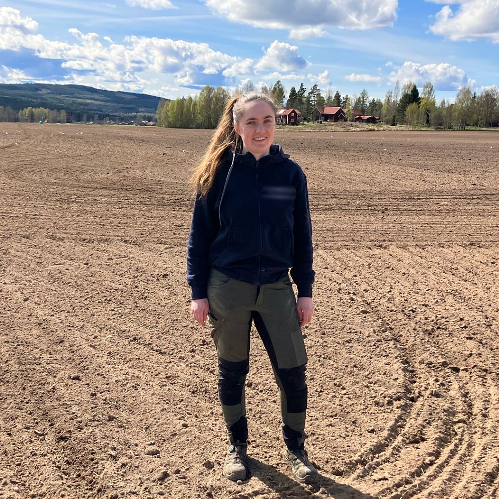 En kvinna står på en öppen åker under blå himmel med moln och skog i bakgrunden.