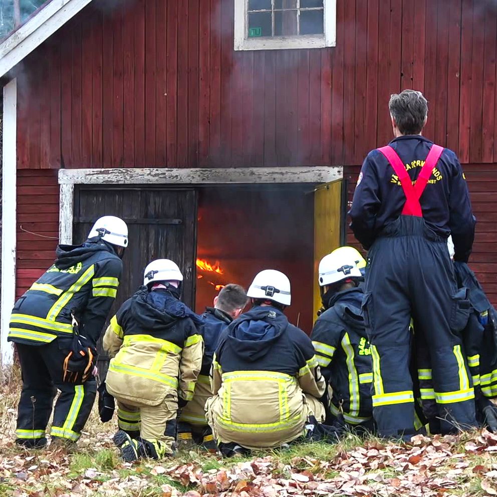 En grupp brandmän i skyddskläder observerar en brand i en röd stuga. Rök stiger från byggnaden, och de är placerade på knä nära en dörröppning för att bedöma situationen.