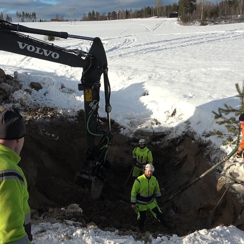 En grävmaskin vid ett hål där det står två varselklädda män