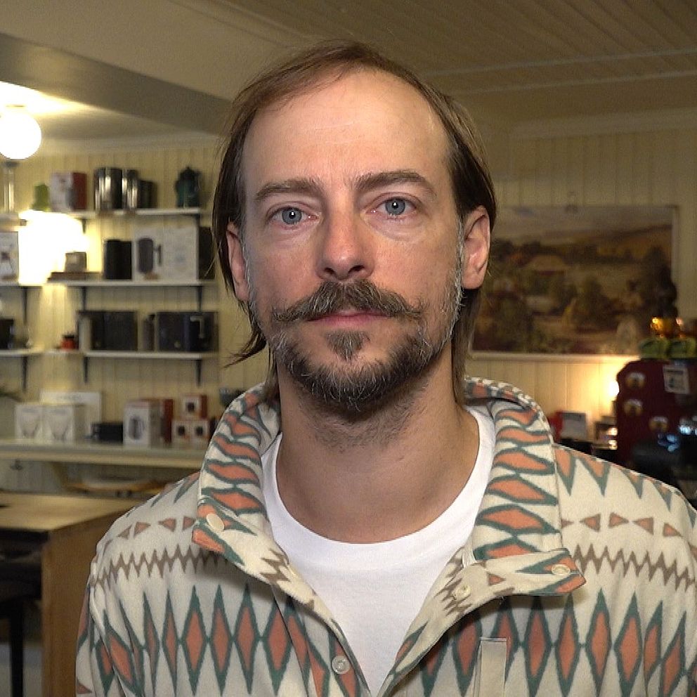 Marcus Izzo har en mönstrad jacka och står i en butik eller café med en ljus och trivsam inredning. Rader av produkter kan ses bakom personen, som verkar vara inriktad på att sälja olika varor. Belysningen är mjuk och skapar en hemtrevlig atmosfär.