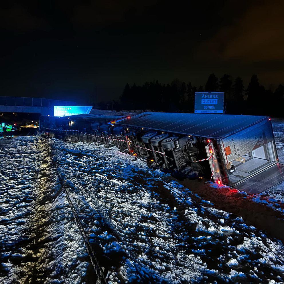 En lastbil har vält på en snöig väg bredvid en motorvägsbro under natten. Räddningspersonal, upplysta av blått blinkande ljus, arbetar vid platsen i bakgrunden.