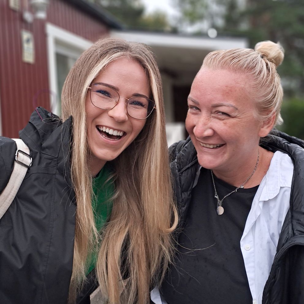 Två kvinnor står arm i arm utanför rött hus i Ronna och skrattar till kameran