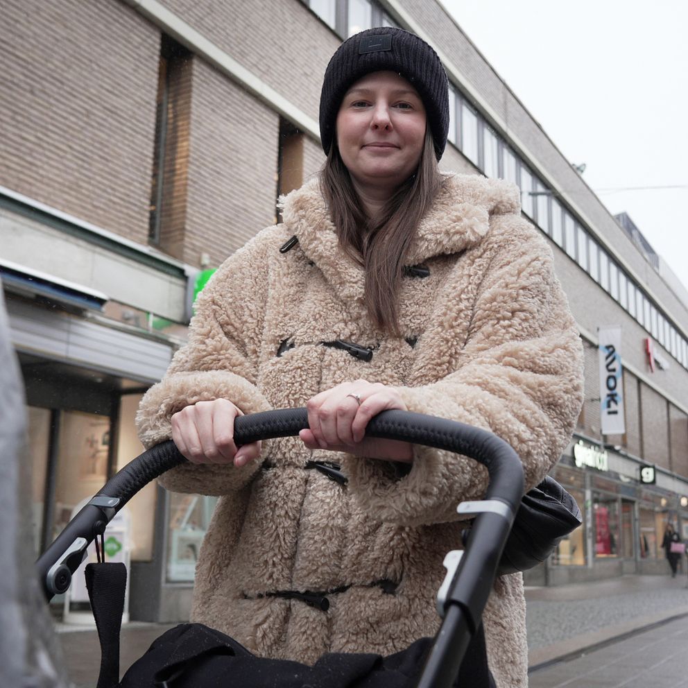En person i en beige teddyjacka står och skjuter en barnvagn längs en gata i en stadsmiljö. I bakgrunden syns kommersiella byggnader och butiker. Vädret är grått och disigt, vilket tyder på en kall dag.