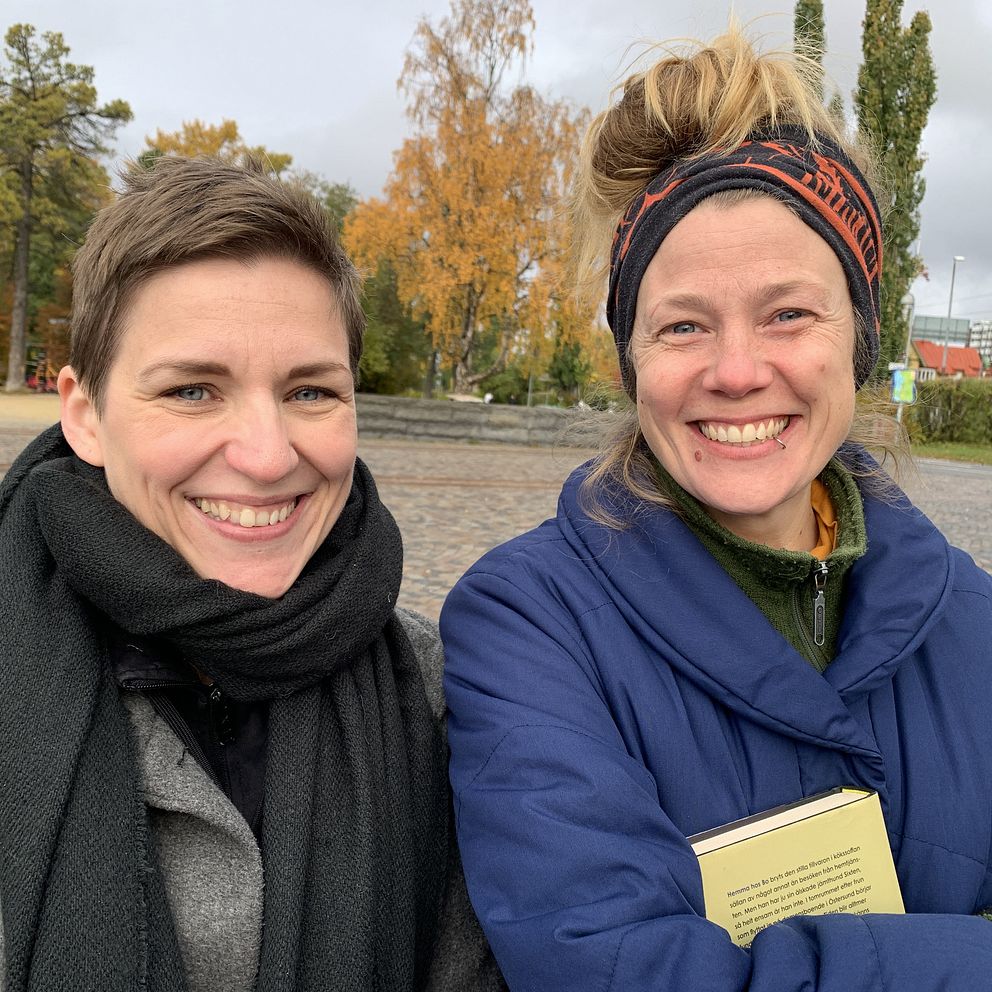 Två personer står nära varandra i en park med höstträd i bakgrunden. Den ena personen har på sig en svart scarf och en grå jacka, medan den andra har en blå jacka och en färgglad huvudbonad. De verkar vara ute och njuter av det kyliga vädret. En bok hålls i den andra personens hand.