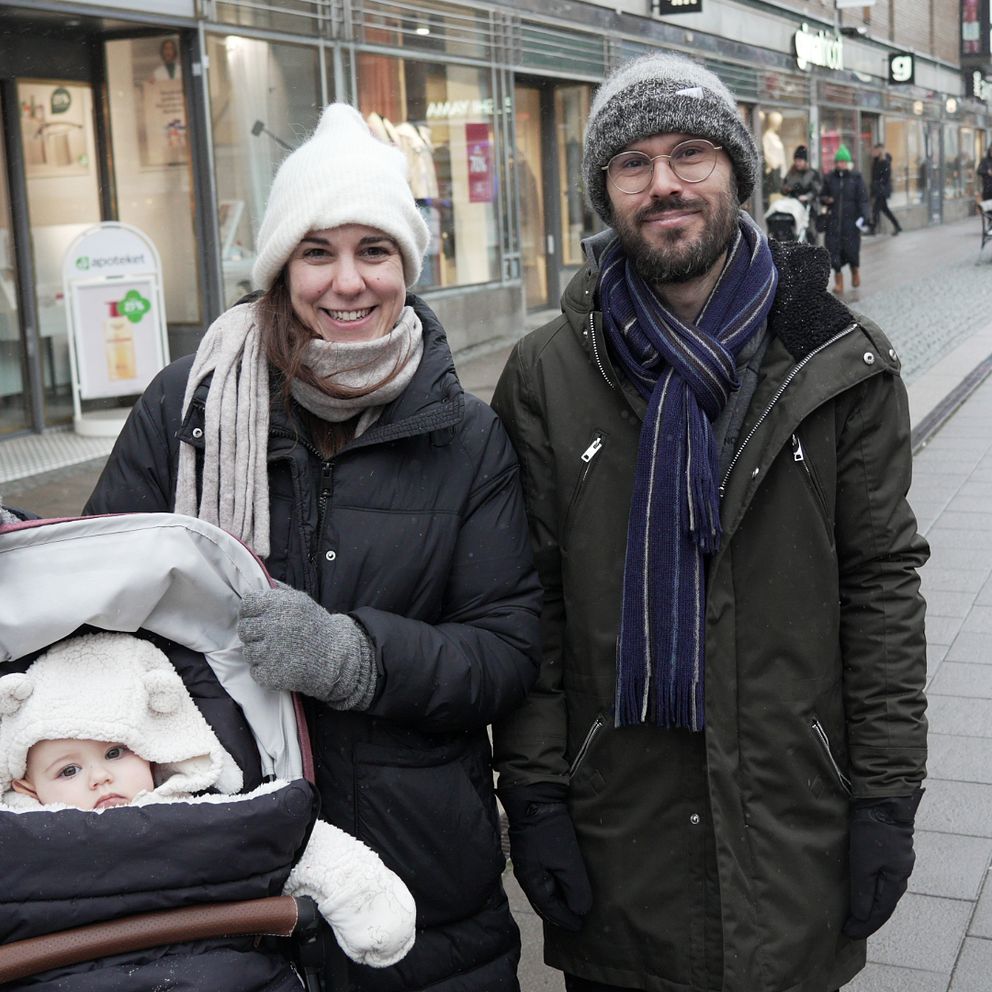 En vuxen kvinna och en man står på en gågata i en stad, iklädda vinterkläder, med en barnvagn framför dem. Barnet i vagnen är insvept i en vit filt. I bakgrunden syns andra människor som promenerar samt butiker längs gatan.