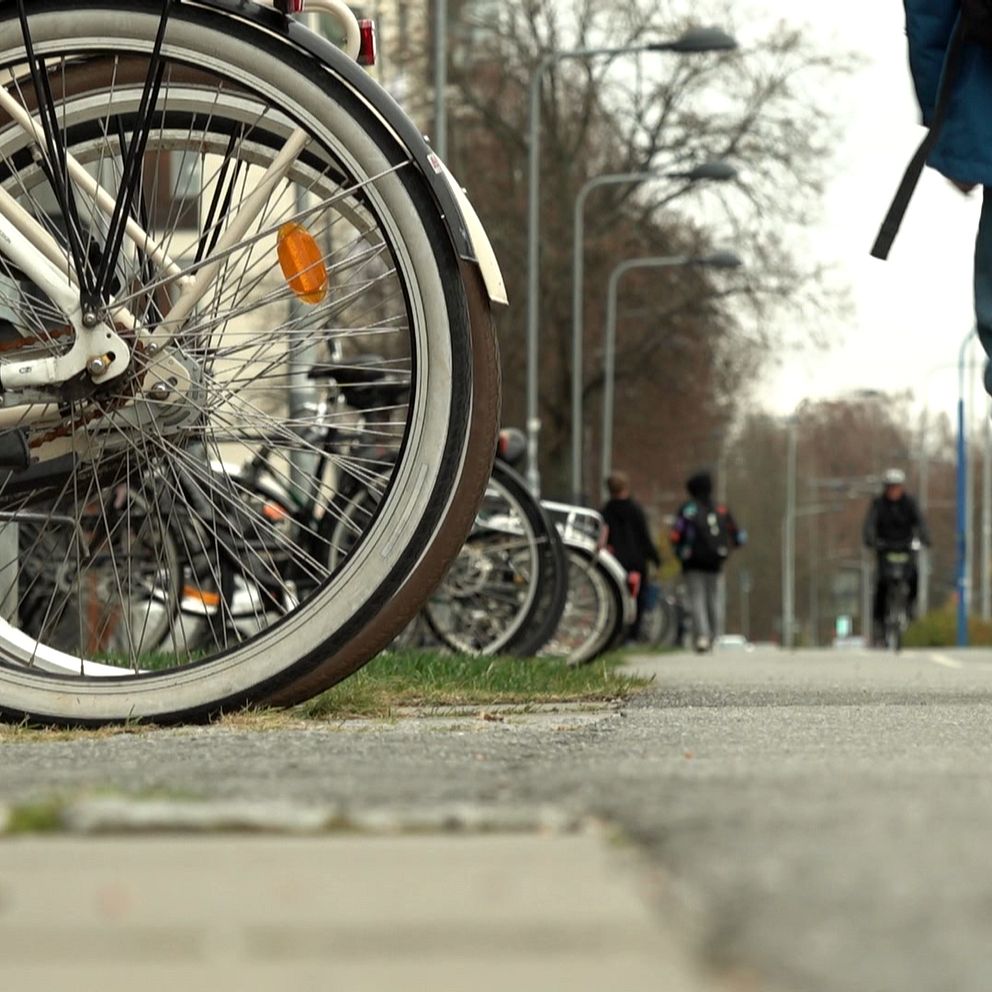 Cyklar står parkerade längs en trottoar medan en person går förbi. Fokus är på en cykel med en stor bakhjul och fälgar, medan andra cyklar kan skönjas i bakgrunden. Scenen skildrar en livlig stadsmiljö med cyklister och fotgängare.