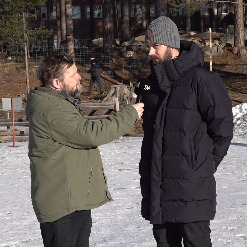 En reporter håller en mikrofon och intervjuar en person iförd en svart vinterrock och grå mössa på en snötäckt yta utomhus.