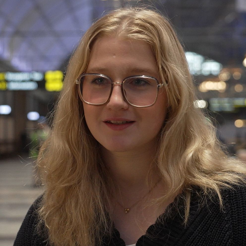 En kvinna med långt blont hår står i en livlig inomhusmiljö, troligen en järnvägs- eller flygterminal. Bakgrunden inkluderar skyltar och människor i rörelse.