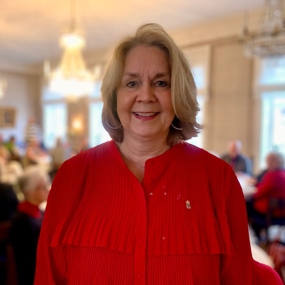 En person i en röd blus står i förgrunden av en social samling, där många andra personer är synliga i bakgrunden i en festlig miljö. Rummet har ljusa väggar och är inrett med kristallkronor och fönster som släpper in naturligt ljus. De andra deltagarna verkar vara engagerade i samtal eller aktiviteter.