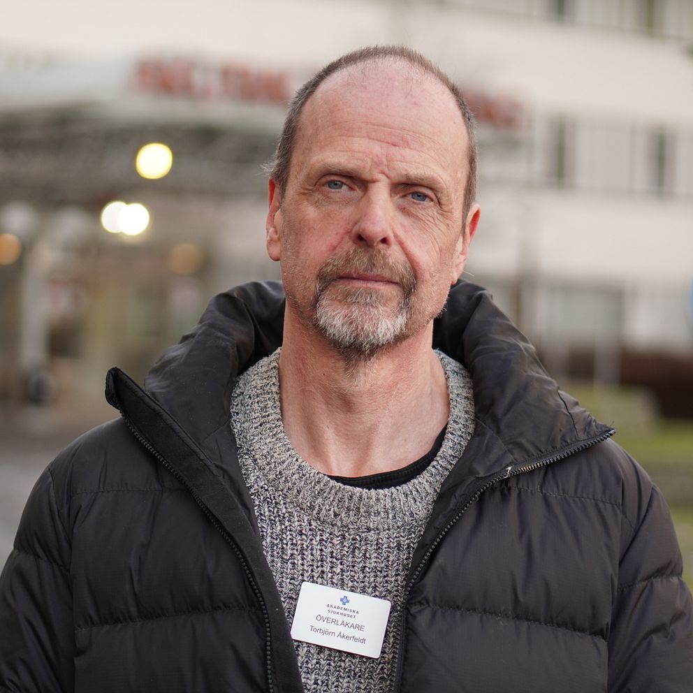 En person i en svart jacka och en grå tröja står framför en byggnad med en skylt. Personen bär ett namnskylt på bröstet. I bakgrunden syns en parkeringsskylt och träd.