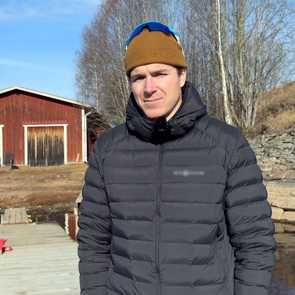 Person står på brygga framför röd byggnad med garageportar, skogsbeklädda kullar i bakgrunden.