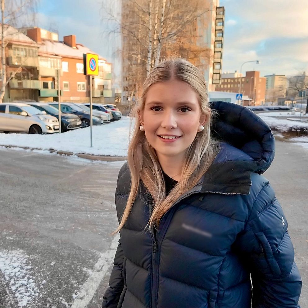 En person klädd i en mörk vinterjacka står dock i en snötäckt stadsmiljö. Bakgrunden visar byggnader och parkering, med några bilar synliga. Det är klart väder med en svag sol som lyser över landskapet.