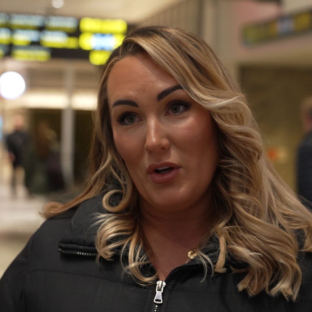 En person står inomhus på en flygplats, omgiven av andra resenärer och med informationsskyltar synliga i bakgrunden.