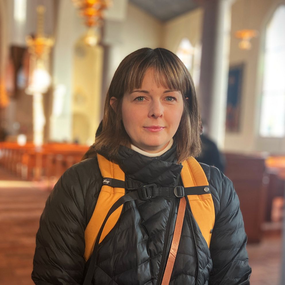 Christina Jardenius, står med svart dunjacka och gul ryggsäck i mittgången inne i stora kyrkan i Östersund.