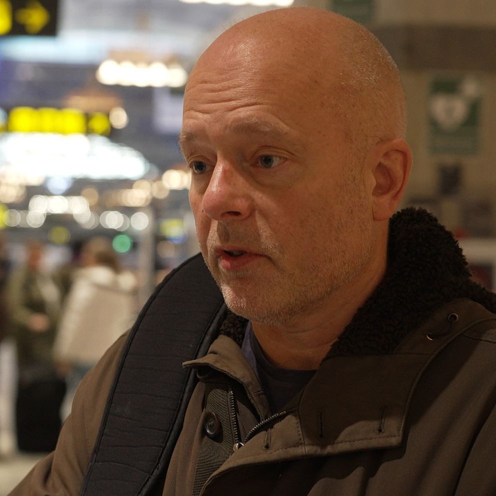 En man i vinterkläder står på en livlig tågstation med människor i bakgrunden som går förbi och informationstavlor som visar tågtider.