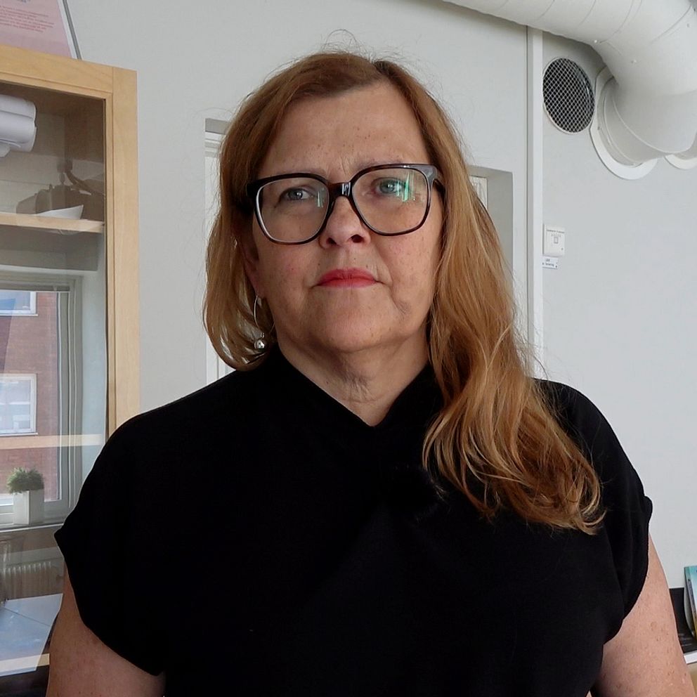 En kvinna med långt brunt utslaget hår som faller över vänster axel, hon har svarta glasögon och svart v-ringad tröja som går högt upp mot halsen.