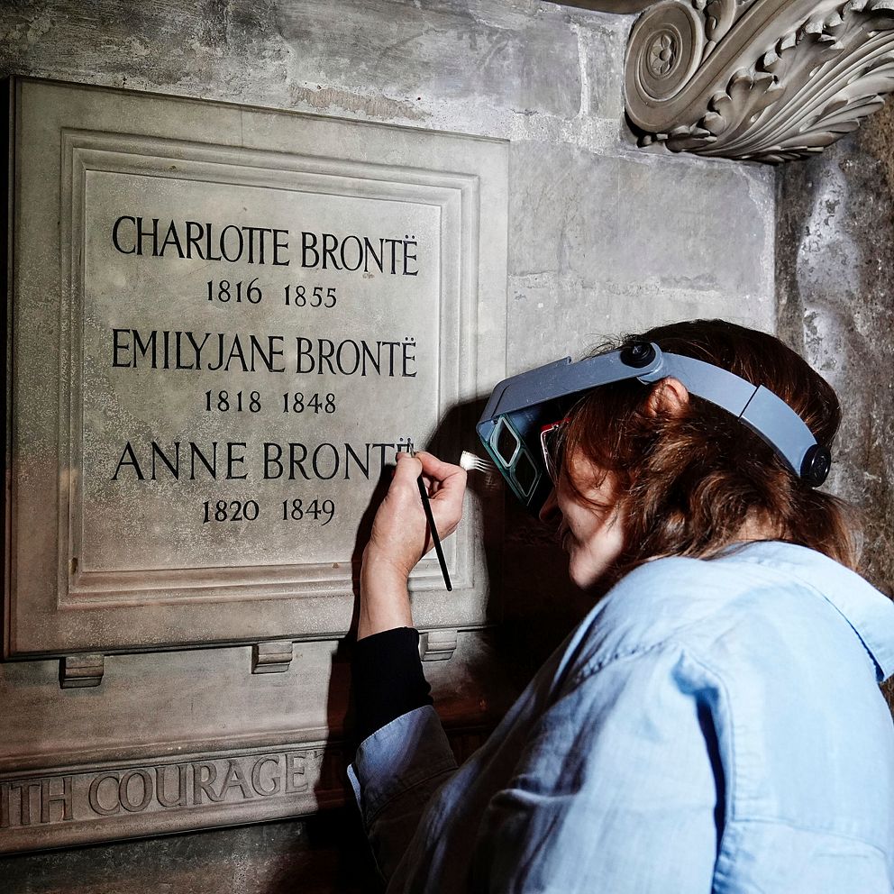 En person med skyddsglasögon arbetar med att stava rätt minnesstenen för författarna Charlotte, Emily och Anne Brontë, med årtal angivna för varje författare.