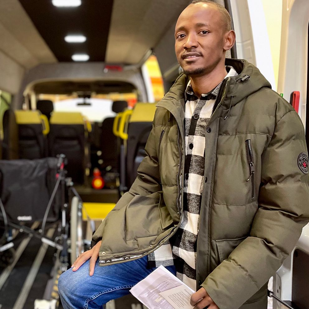 En person sitter i en ambulans med ett rullstolsskåp i bakgrunden, omgiven av gula säten och interiörbelysning. Personen håller i ett dokument och bär en grön jacka med en randig skjorta.