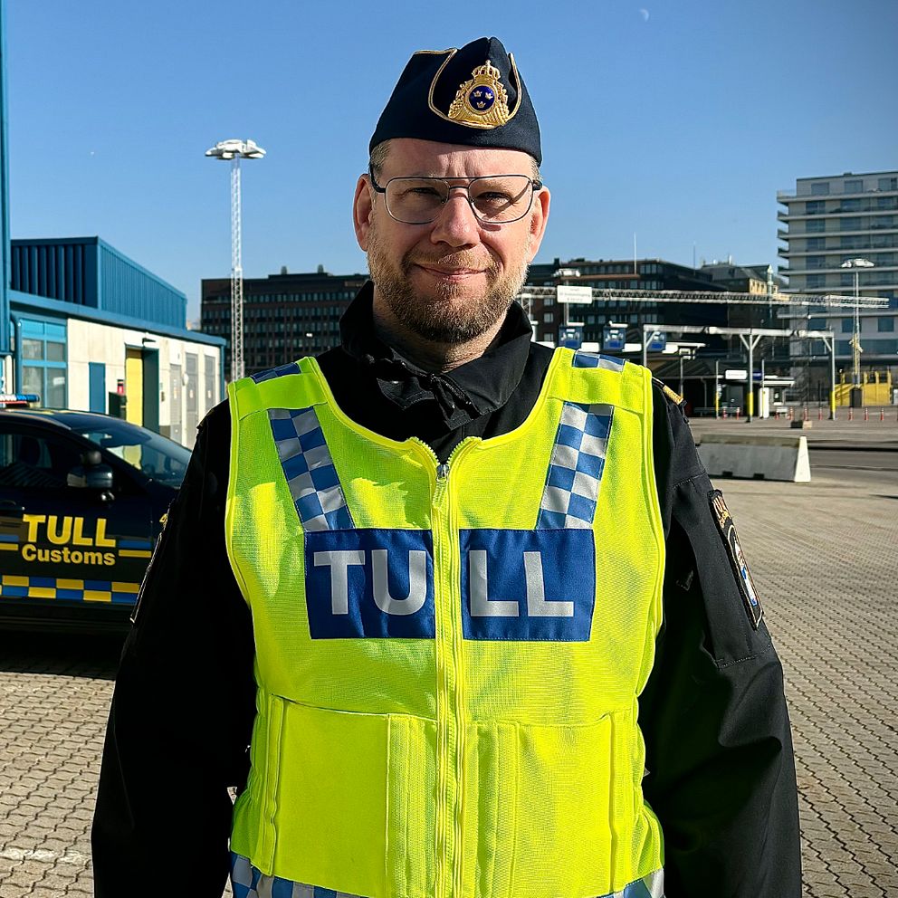 Tulltjänsteman i uniform står framför en tullbil vid en hamnmiljö.