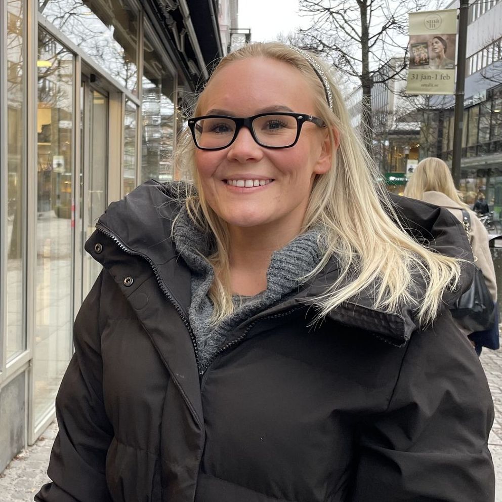 En kvinna i en svart vinterjacka står på en trottoar i en stadsmiljö med glasfasader och butiker på ena sidan. I bakgrunden syns människor som går längs gatan, samt träd och gatubelysning.