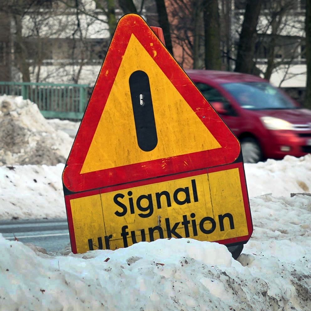 Varningstriangeln i gult och rött med texten ”Signal ur funktion” står lutad vid vägkanten, omgiven av snöiga förhållanden medan bilar passerar i bakgrunden.