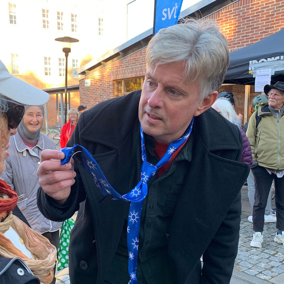 Henrik Kruusval i en svart jacka visar en medalj med blått band för en annan person som bär en solhatt och en scarf. I bakgrunden syns flera personer samt ett tak med en skylt som kan föreslå ett evenemang eller en marknad.