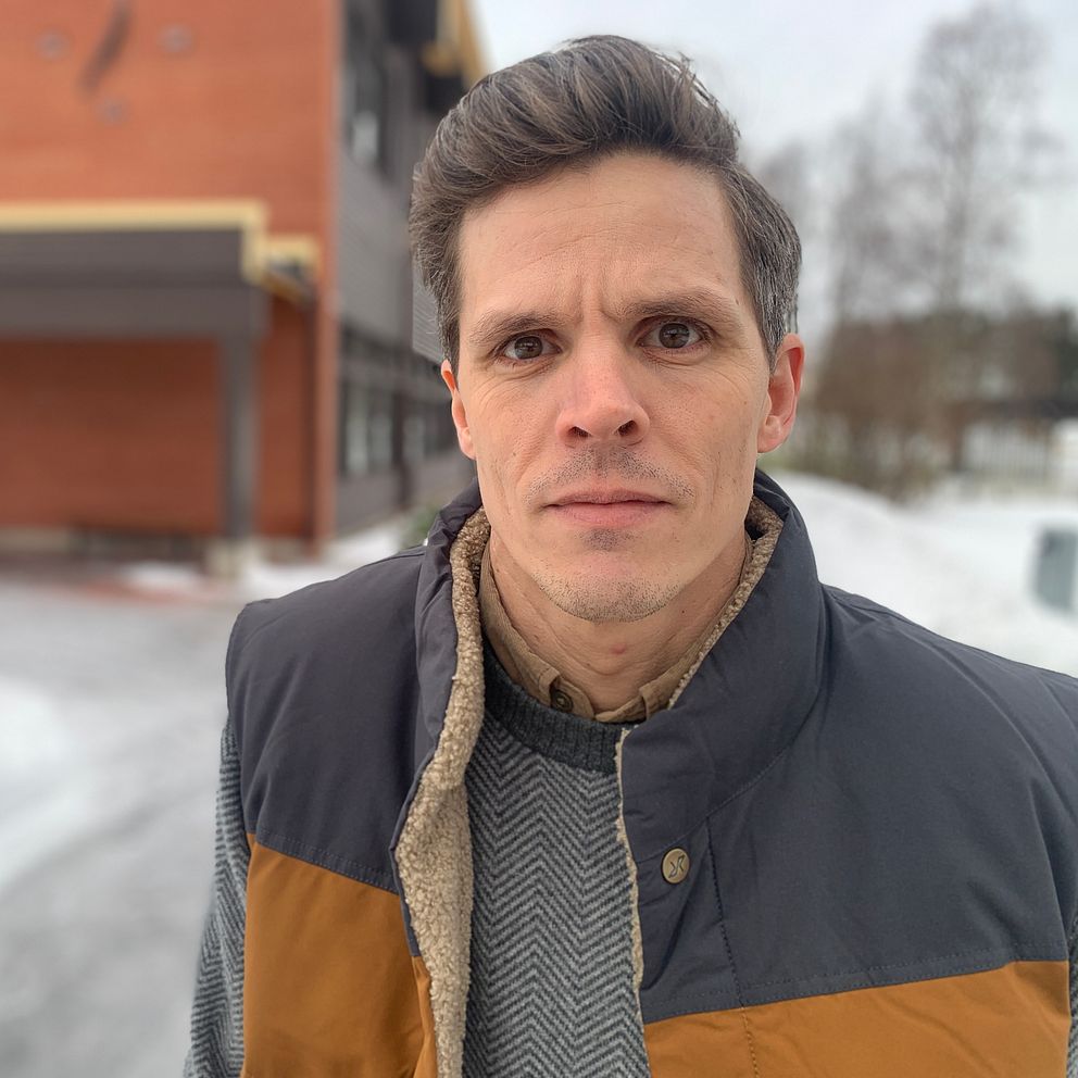 En person med mörkt hår står utomhus framför en tegelbyggnad på vintern. Marken är täckt av snö och några träd syns i bakgrunden. Personen bär en vinterjacka med bruna och svarta färgade sektioner.