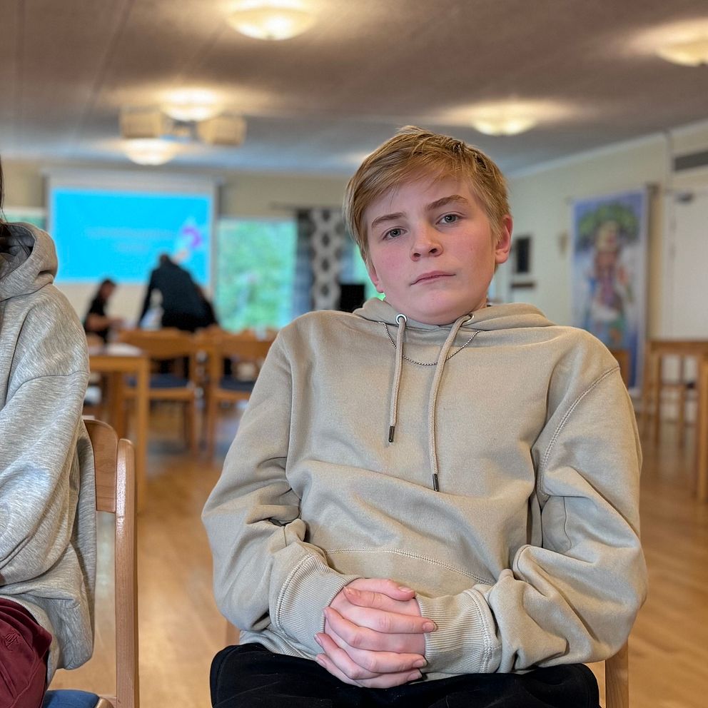 Två barn sitter på stolar i en inomhusmiljö, klädda i grå hoodies. Rummet har träbord och stolar med en stor projektor i bakgrunden som visar en bild. Belysningen är mjuk och rummet har en avspänd atmosfär.