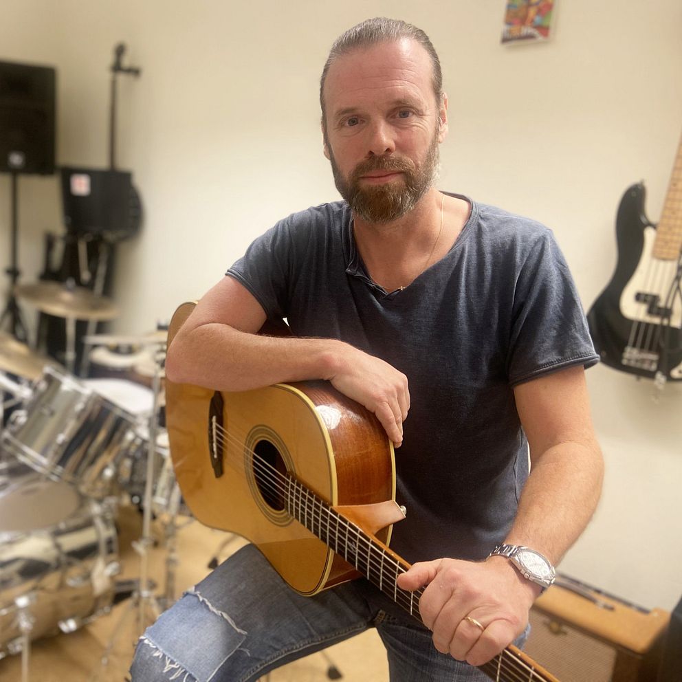 Mattias Hultman sitter med en akustisk gitarr i knät i en musikstudio, med ett trumset synligt i bakgrunden.