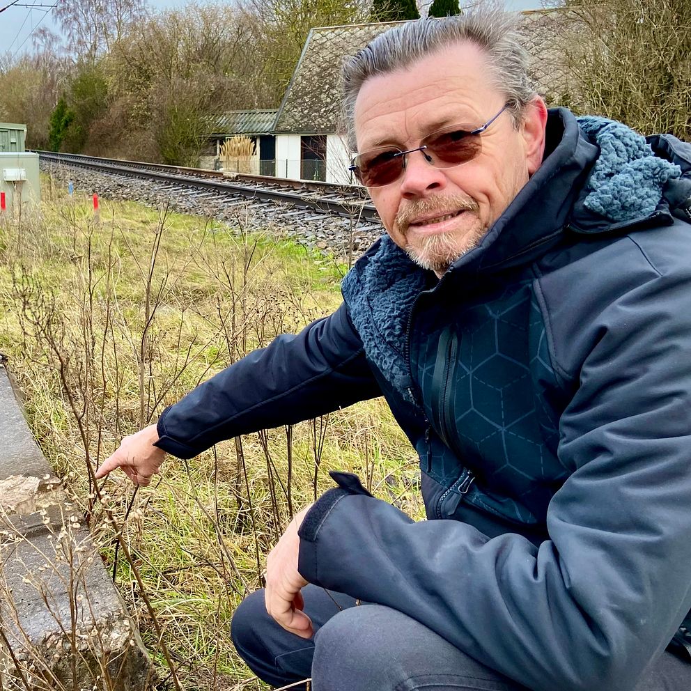 En person i mörk jacka pekar mot en del av ett defekt järnvägsspår som ligger vid sidan av spåret i en lantlig miljö. I bakgrunden syns ett stängsel, järnvägsspår och en byggnad.