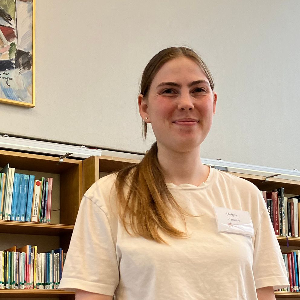 En ung kvinna står framför en bokhylla i en biblioteksmiljö. I bakgrunden syns en färgglad målning som hänger på väggen. Bokhyllan är fylld med böcker i olika färger och storlekar.