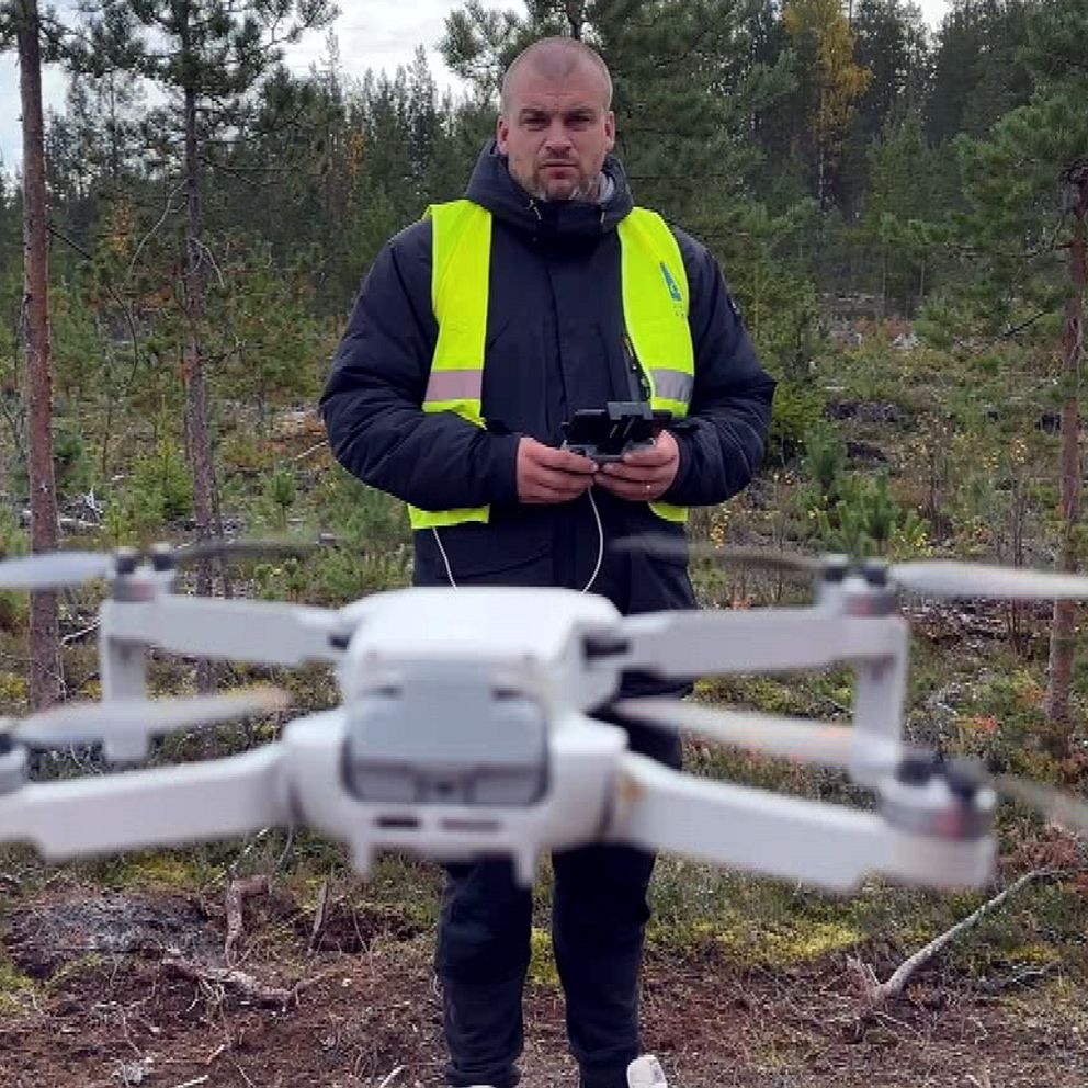 Bild på en drönare i luften och drönarens pilot i en skog i Örnsköldsvik i bakgrunden.