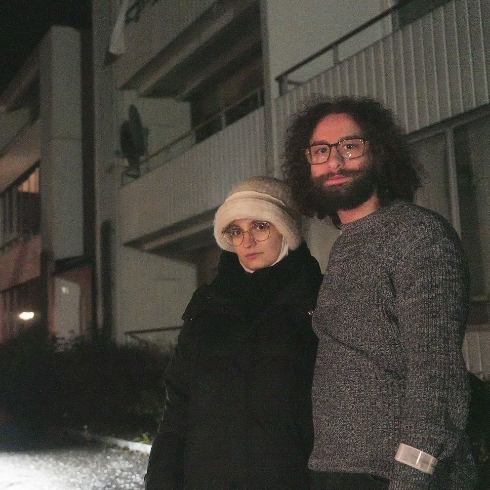 Två personer står utomhus framför en byggnad under natten, med en bil som har strålkastarna på i bakgrunden. Byggnaden har flera fönster, varav några är upplysta, och det finns gröna växter vid marken.