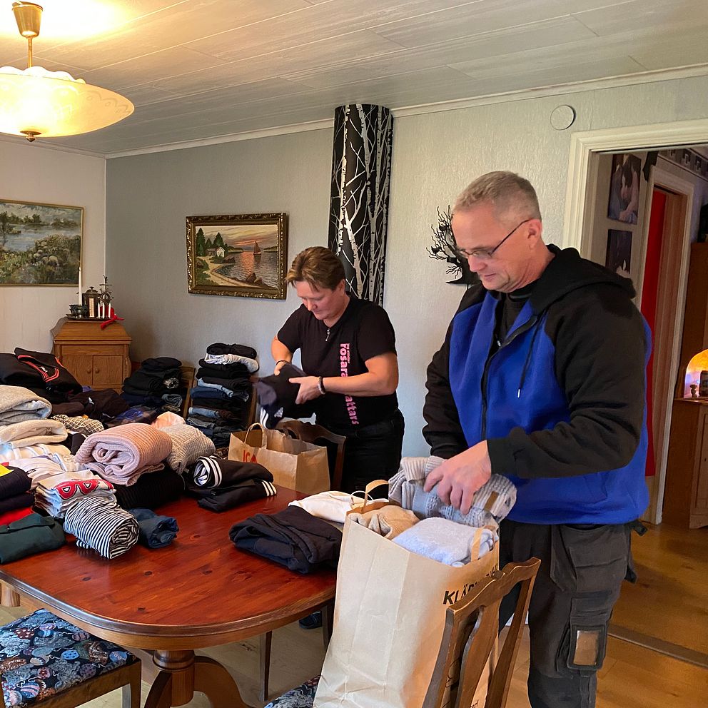 Två personer packar kläder på ett bord i ett vardagsrum. Bordet är belamrat med vikta kläder av olika slag, och det finns papperskassar som används för förvaring. Rummet har en hemtrevlig inredning med en lampa i taket och väggkonst.