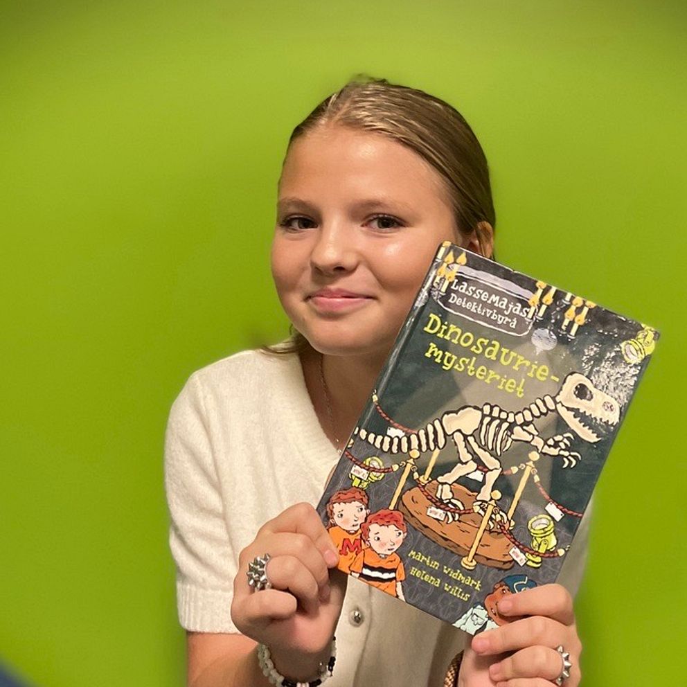 En person håller upp en bok med titeln ”Dinosaurier-mysterier” i framkant, med en färgglad bakgrund i grön nyans. Boken innehåller en illustration av ett dinosaurieskelett och två barn på omslaget.