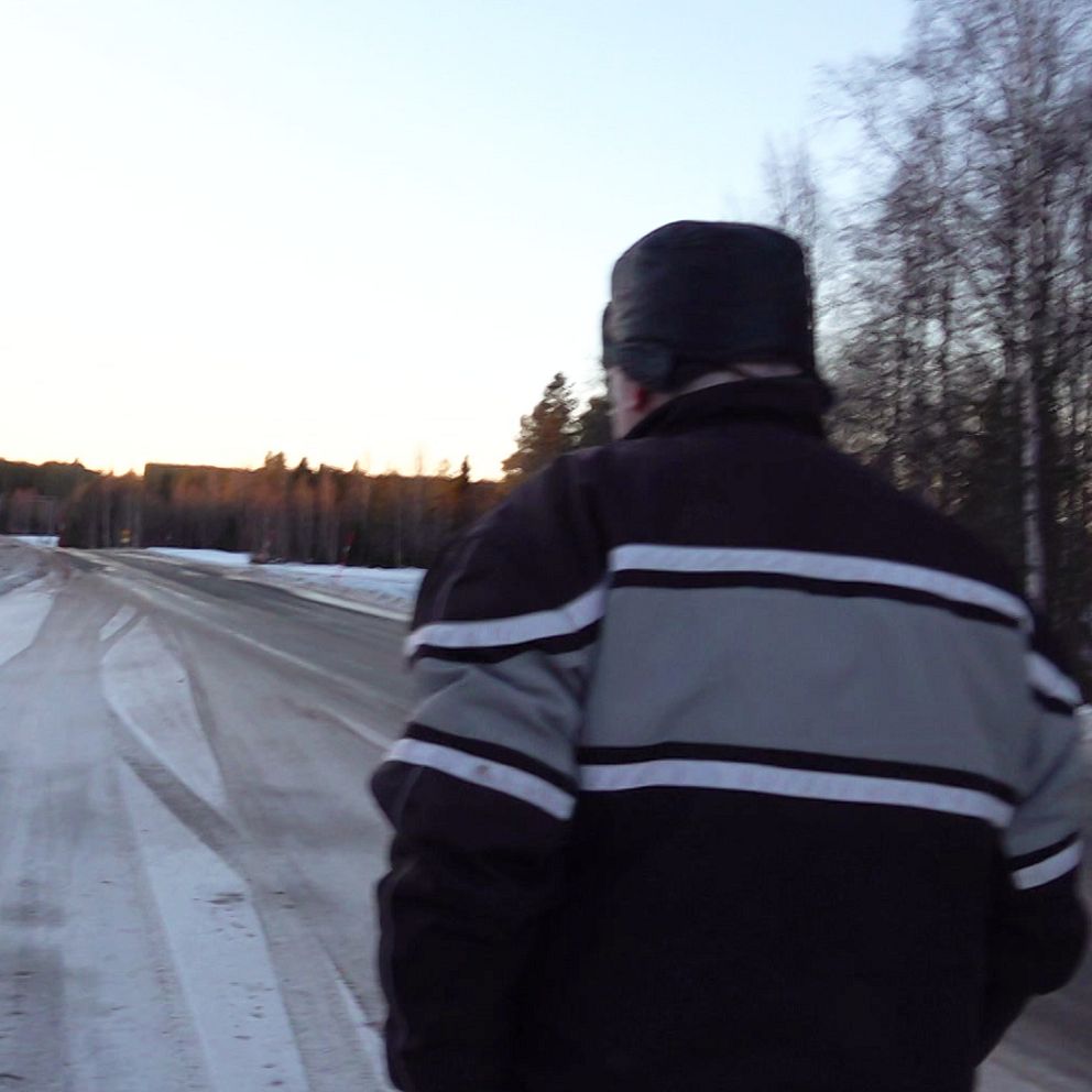 En person går längs en snöig väg med ett trädområde på sidan. Vägen är delvis belagd med snö, och i bakgrunden syns en solnedgång som färgar himlen. Det finns några vägskyltar i närheten.