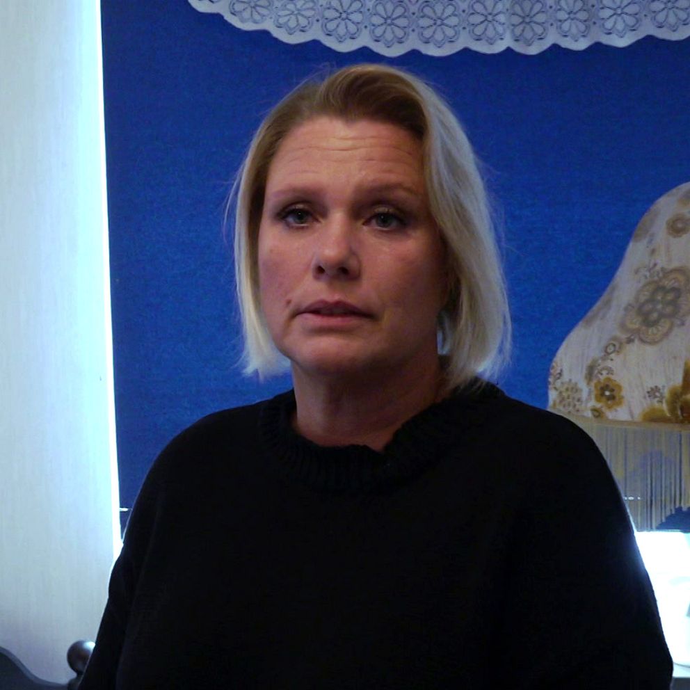 En person står i ett rum med en ljus vägg och en mörk säng i bakgrunden. En bordslampa syns till höger, och en blå gardin med mönster hänger i fönstret. Äldreomsorg, hemtjänst, missförhållande.