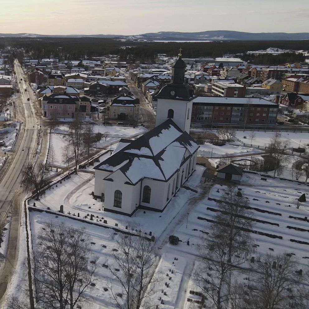 Utsikt över en snöklädd Sveg med en kyrka i förgrunden. Gatorna är tomma och omgivande byggnader syns i bakgrunden, inklusive moderna kontorshus. Landskapet är präglat av snö och vinterljus, vilket skapar en lugn atmosfär.