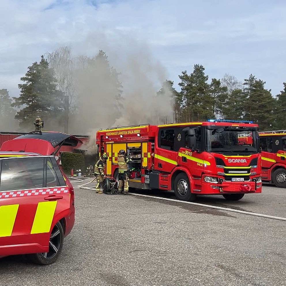 Brandbilar vid brandplats, rök stiger från byggnaden bakom dem.