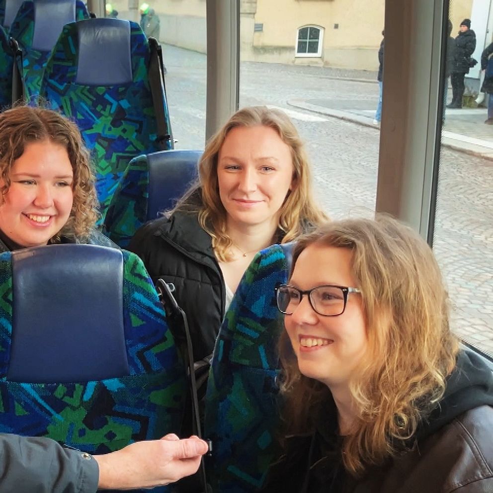En person står i en buss och pratar med tre passagerare som sitter på sätena, medan bussens interiör med mönstrade säten och fönster i bakgrunden syns.