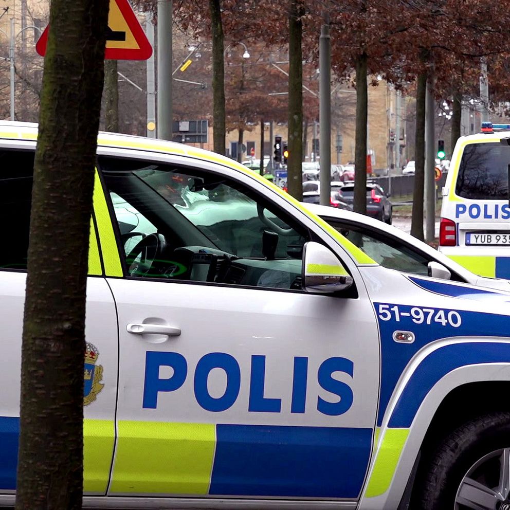Två poliser i uniformer går framåt efter att tre personer blivit knivskurna vid en frisörsalong på Mölndalsvägen i centrala Göteborg. En av dem håller i en brun papperspåse. Till höger finns en polisbil parkerad, med tydlig text som visar att det är en polisfordon. I bakgrunden syns träd och byggnader som skapar en stadsmiljö.