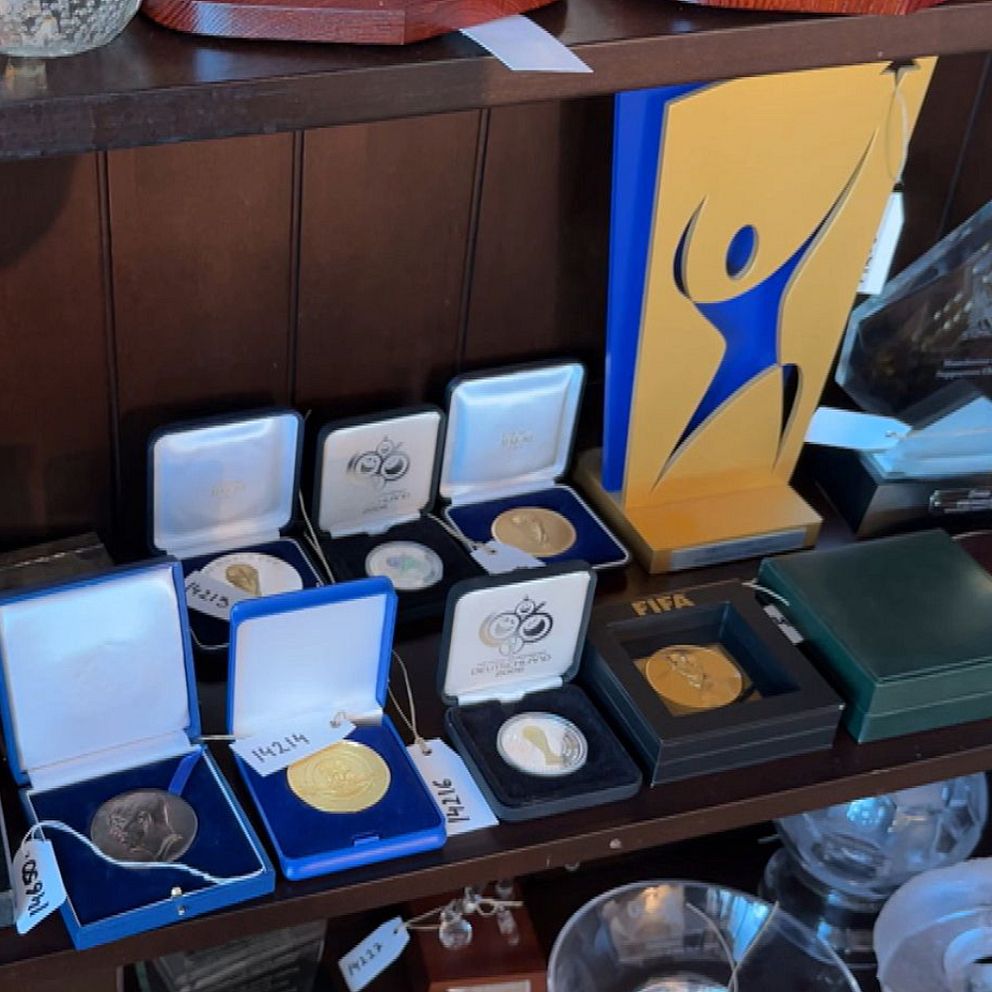 På vänstra sidan av bilden håller en person upp en papperslista framför en orange jacka med en engelsk fotbollströja. På högra sidan visas en hylla med olika medaljer och utmärkelser, inklusive insatta mynt och en platta med en stiliserad figur i blått och gult.