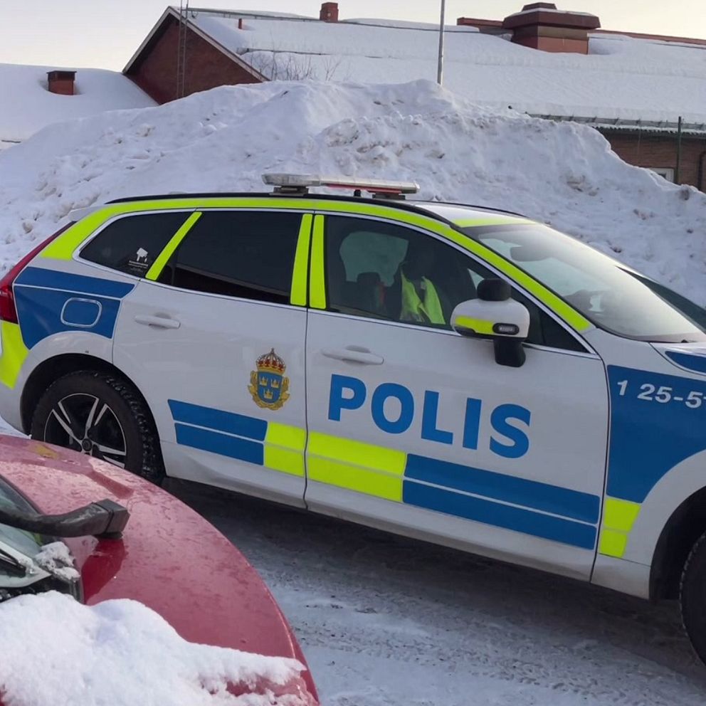 En polisbil står parkerad på en snöig gata framför en hög med snö. Tre poliser står och samtalar nära en byggnad i bakgrunden. Det är vinter, vilket syns på snötäcket på bilarna och marken.