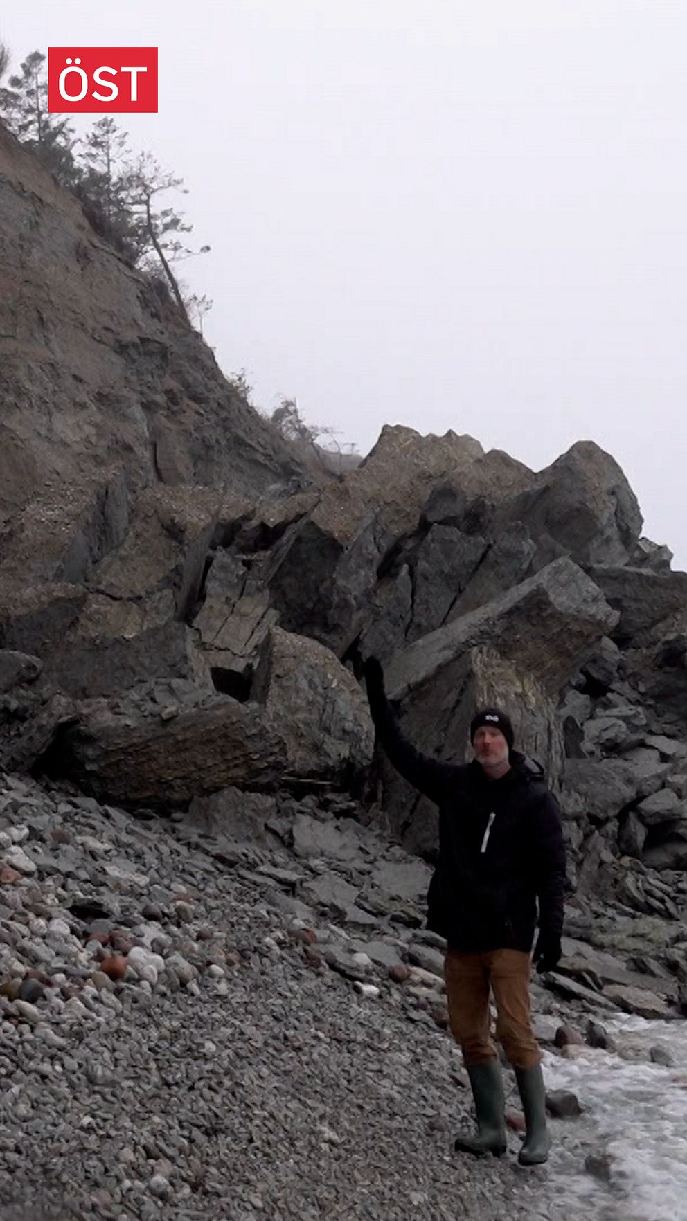 En person står vid en stenig strand på Gotland framför en stor bergsklippa som har kollapsat, med stenar och klippblock utspridda.