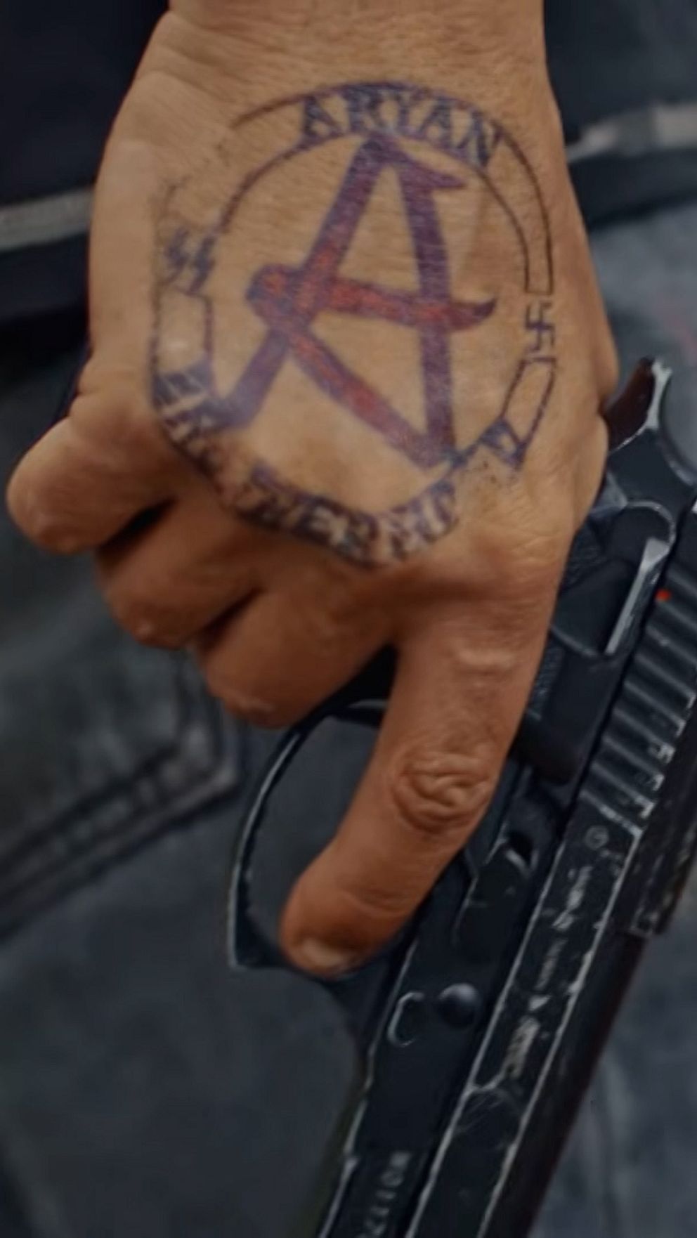 En man håller en pistol i handen med en stor tatuering föreställande en bokstav ”A”.