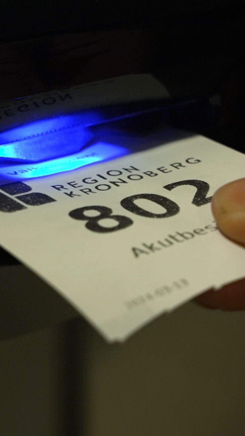 En hand som sitter fast vid en papperslapp med numret 802, som placeras i en dispenser med blå ljus, märkt med ”Region Kronoberg” och texten ”Akutbesök”.