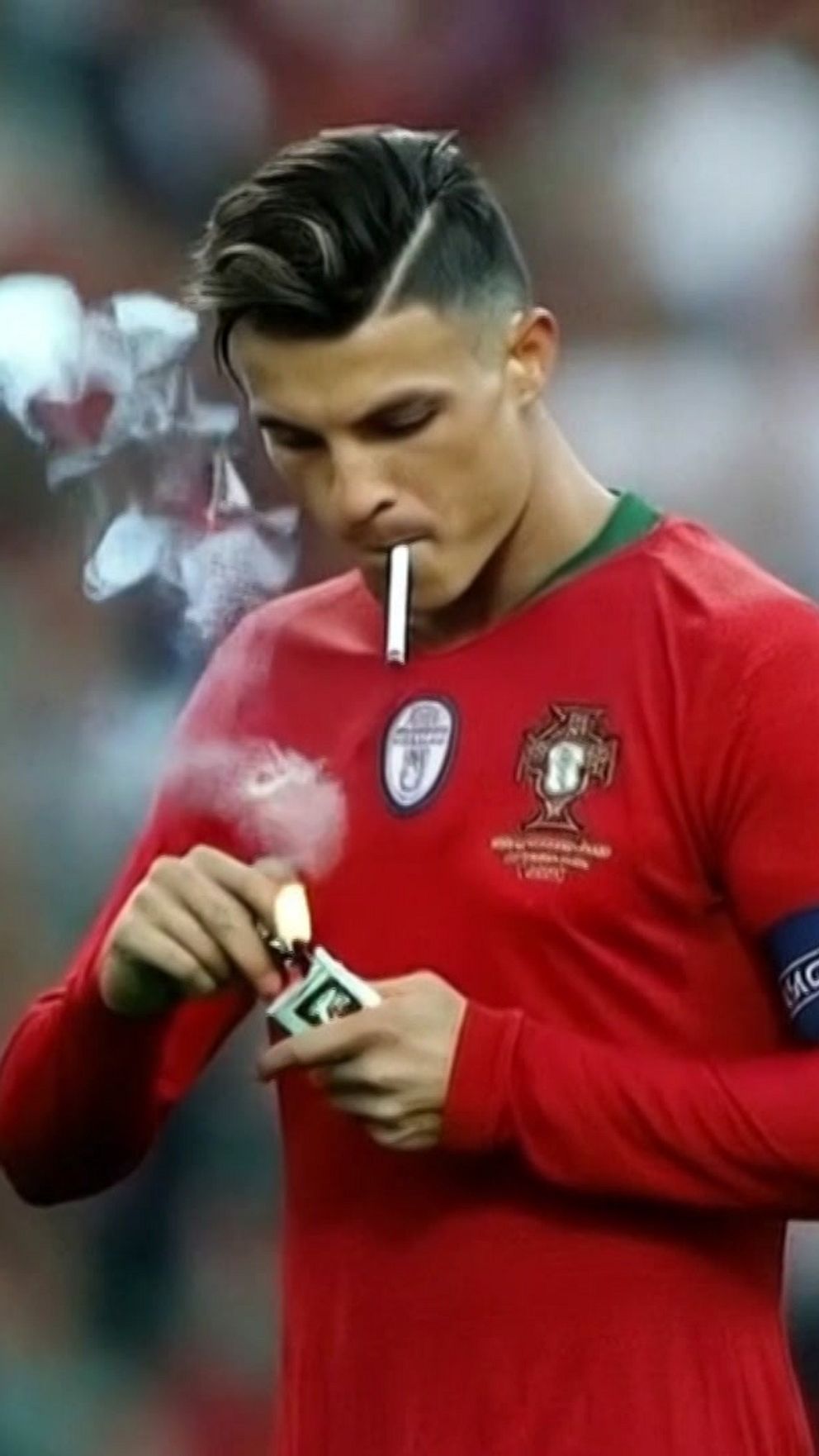 En fotbollsspelare i röd tröja med Portugals flagga och kaptens-armbindel ser ut att tända en cigarett med en tändsticka. Han har cigaretten i munnen.